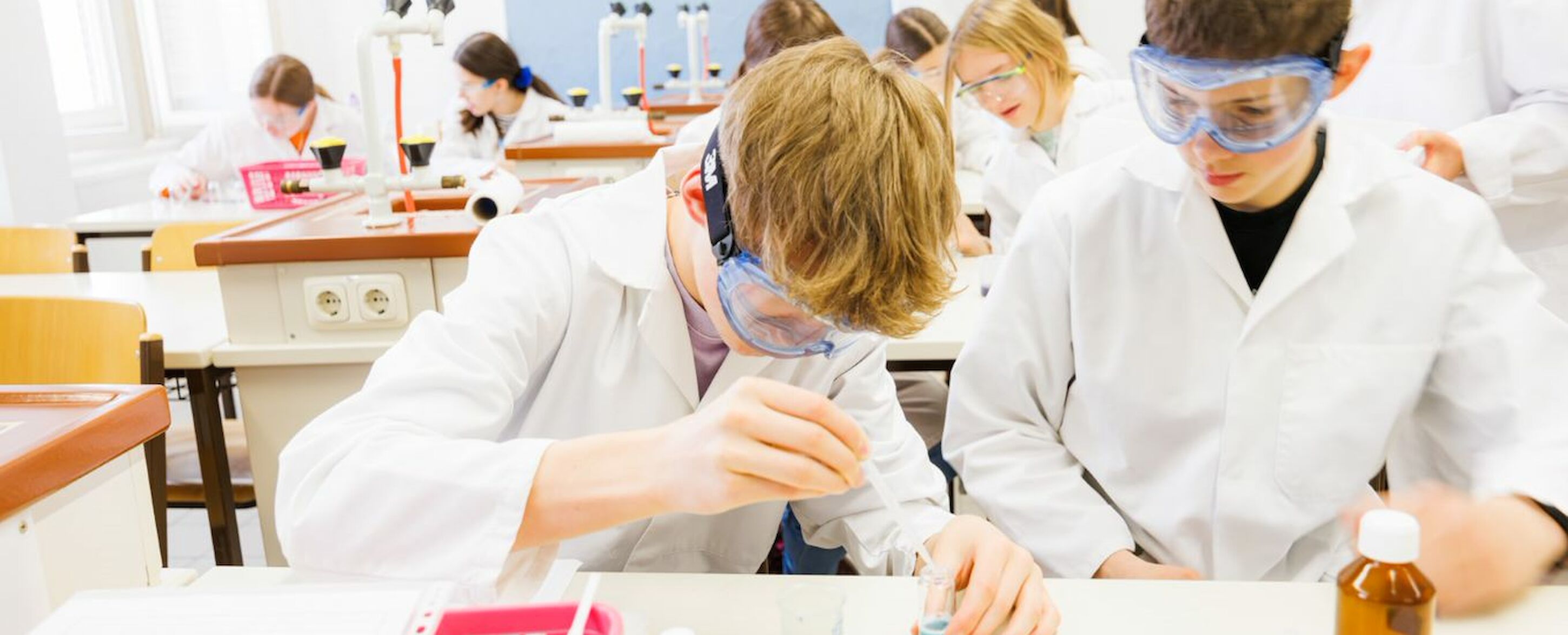 Schüler in Laborkitteln und mit Schutzbrillen führen in einem naturwissenschaftlichen Klassenzimmer Experimente durch.