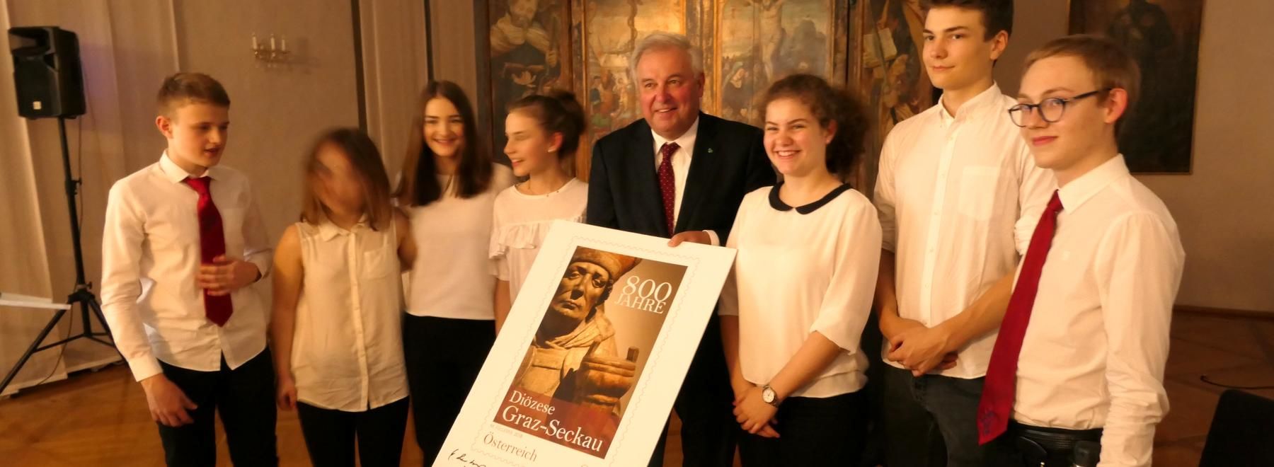 Sieben Personen posieren in einem Innenraum mit einer großen Plakette, die eine Statue und einen Text zeigt.