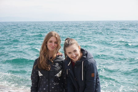 Zwei junge Frauen, die an einem bewölkten Tag am Meer stehen und lächeln.