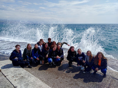 Eine Gruppe Jugendlicher posiert am Meer, während hinter ihnen an einem sonnigen Tag die Wellen schlagen.