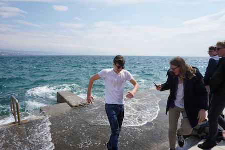 Ein junger Mann in nassen Kleidern kommt aus dem Meer, während Freunde zusehen und lachen.