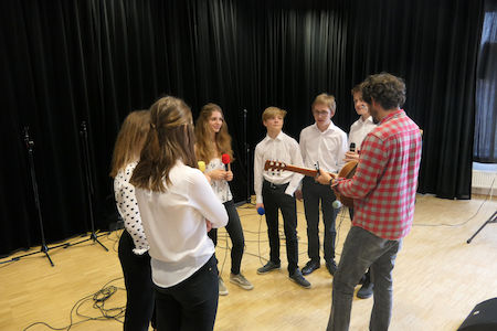 Eine Gruppe von Schülern singt, während ein Mann in einem Proberaum Gitarre spielt.