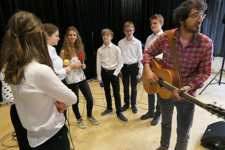 Eine Gruppe Jugendlicher singt mit einem Mann, der in einem Musikstudio Gitarre spielt.