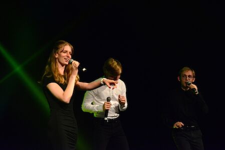 Drei Personen, die auf der Bühne singen und jeweils ein Mikrofon in der Hand halten, vor einem dunklen Hintergrund.