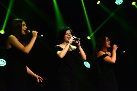 Drei Frauen in schwarzen Kleidern singen auf der Bühne unter grünen Scheinwerfern.