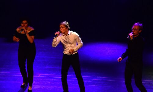 Drei Personen singen auf der Bühne bei violettem Licht.