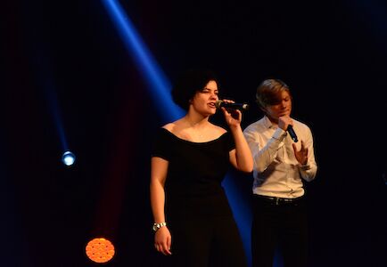 Zwei Personen singen auf einer dunklen Bühne mit Scheinwerfern hinter ihnen in Mikrofone.