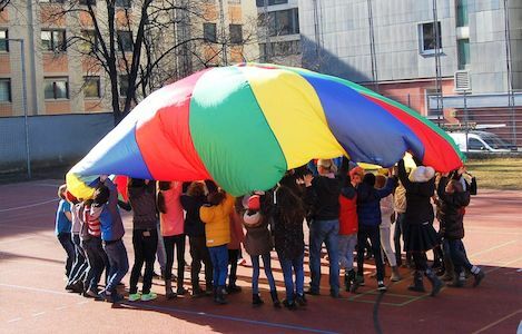 Kinder heben gemeinsam einen bunten Fallschirm auf einem Spielplatz im Freien auf.