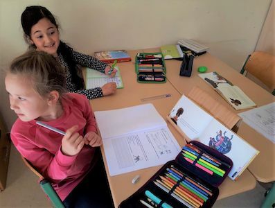 Zwei Mädchen sitzen an einem Schreibtisch mit aufgeschlagenen Notizbüchern und bunten Stiften und arbeiten zusammen.