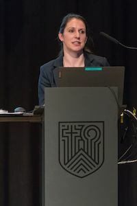 Frau, die an einem Podium mit einem Wappen spricht und einen Vortrag hält.