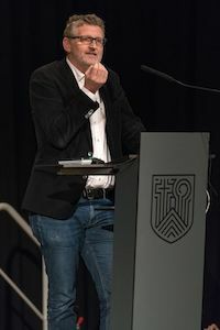 Ein Mann in einem schwarzen Blazer spricht auf einem Podium auf der Bühne.