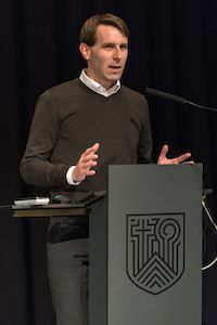 Mann, der an einem Podium mit einem Symbol spricht, trägt einen dunklen Pullover und ein weißes Hemd.