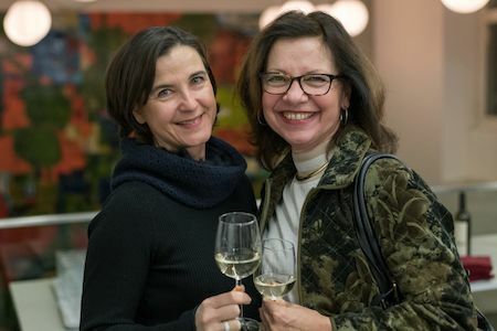 Zwei lächelnde Frauen mit Weißweingläsern bei einer Veranstaltung in einem Gebäude.