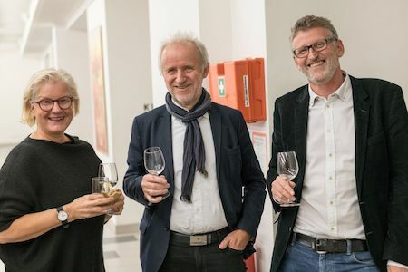 Drei lächelnde Menschen, die bei einer Indoor-Veranstaltung Getränke in der Hand halten und zusammen posieren.
