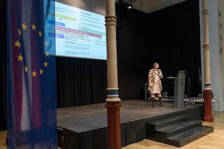Eine Frau spricht auf der Bühne neben einer EU-Flagge und einem großen Bildschirm mit einer Grafik.
