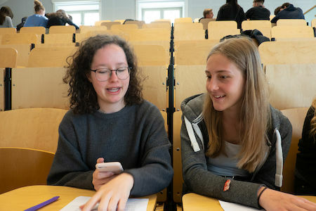 Zwei Studenten sitzen in einem Hörsaal, unterhalten sich und lächeln, während sie Papiere vor sich liegen haben.