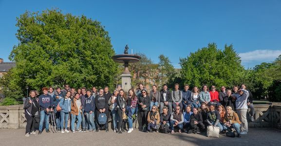 Eine große Gruppe von Menschen posiert gemeinsam vor Bäumen und einem Springbrunnen im Freien.