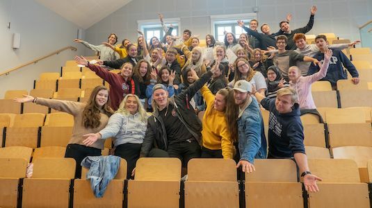 Eine Gruppe von Studenten posiert und lächelt in einem Hörsaal mit ausgestreckten Armen.