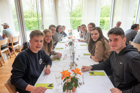 Eine Gruppe junger Leute sitzt lächelnd an einem langen Tisch in einem hellen Restaurant.