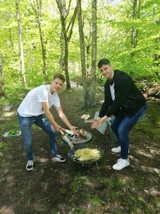 Zwei junge Männer kochen Essen über einem Feuer im Freien in einem Waldgebiet.