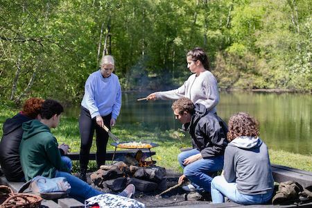 Sechs Personen braten Essen über einem Lagerfeuer in der Nähe eines Sees in einer grünen, bewaldeten Gegend.
