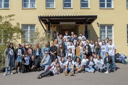 Eine große Gruppe von Menschen posiert gemeinsam vor einem gelben Gebäude an einem sonnigen Tag.