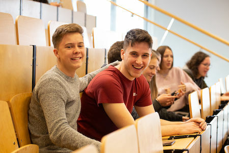 Zwei lächelnde Studenten sitzen in einem Hörsaal mit anderen Studenten im Hintergrund.