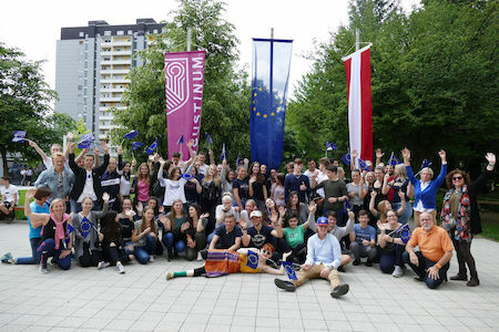 Eine große Gruppe von Menschen posiert im Freien mit Fahnen und Transparenten, winken und lächeln.