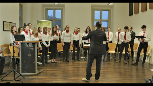 Ein Chor in weißen Hemden und roten Krawatten singt, während ein Dirigent ihn in einen Raum führt.