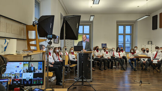 Ein Mann spricht an einem Podium, hinter ihm sitzen maskierte Personen in einem Klassenzimmer.