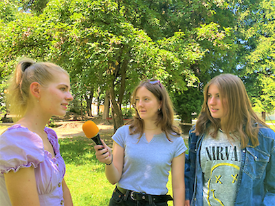 Drei junge Frauen im Freien, eine hält ein Mikrofon und interviewt eine andere.