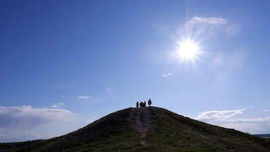 Silhouettierte Figuren stehen auf einer grasbewachsenen Hügelkuppe unter einem hellen, sonnigen Himmel.
