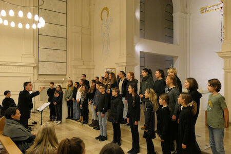 Ein Chor singt in einer Kirche, geleitet von einem Dirigenten, während die Menschen in der Nähe sitzen.