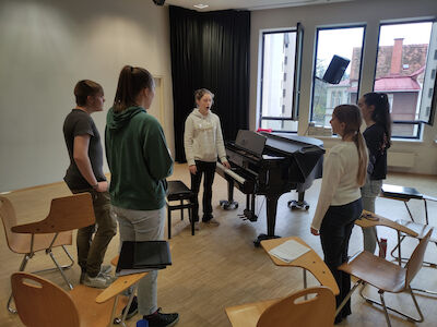 Fünf Jugendliche stehen um ein Klavier in einem Klassenzimmer mit verstreuten Stühlen und offenen Fenstern.