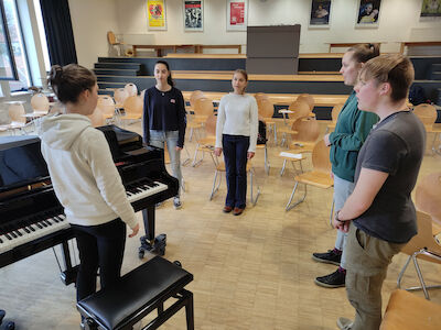 Fünf Schüler stehen in einem Klassenzimmer um ein Klavier herum und bereiten sich auf das Musizieren vor.