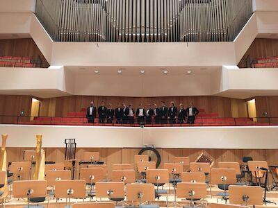 Leere Orchestersitze auf der Bühne, Musiker im Smoking stehen auf dem Balkon dahinter.