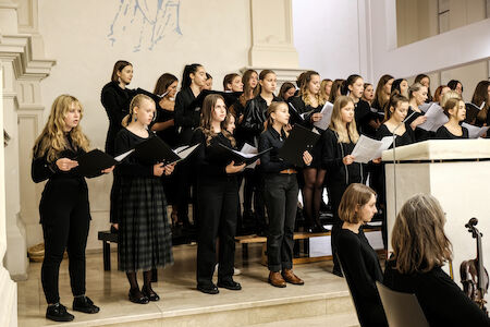 Ein Frauenchor singt in einer Kirche, alle sind schwarz gekleidet und halten Notenblätter in der Hand.
