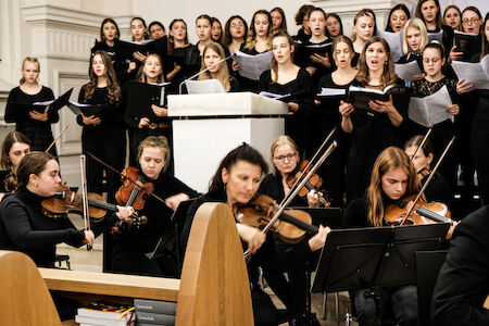 Ein Chor singt und Musiker spielen Geigen bei einer formellen Aufführung in einer Halle.