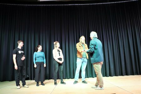 Fünf Personen stehen auf der Bühne vor einem schwarzen Vorhang, zwei schütteln sich die Hände.