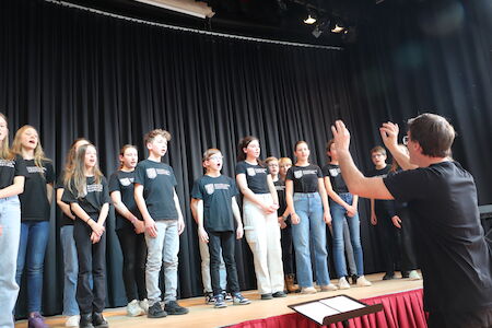 Eine Gruppe von Kindern singt auf der Bühne, während ein Dirigent sie anleitet.