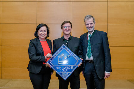 Drei Personen in formeller Kleidung halten ein blaues Schild der Meistersinger Schule.