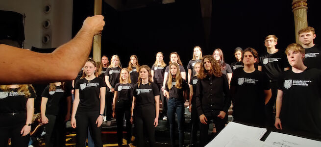 Ein Chor junger Menschen singt auf der Bühne, während ein Dirigent sie leitet.