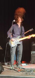 Gitarrist auf der Bühne, headbangend mit verwuscheltem Haar, spielt E-Gitarre.