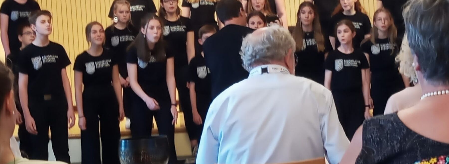 Ein Chor von Jugendlichen singt auf der Bühne, beobachtet von sitzenden Erwachsenen im Vordergrund.
