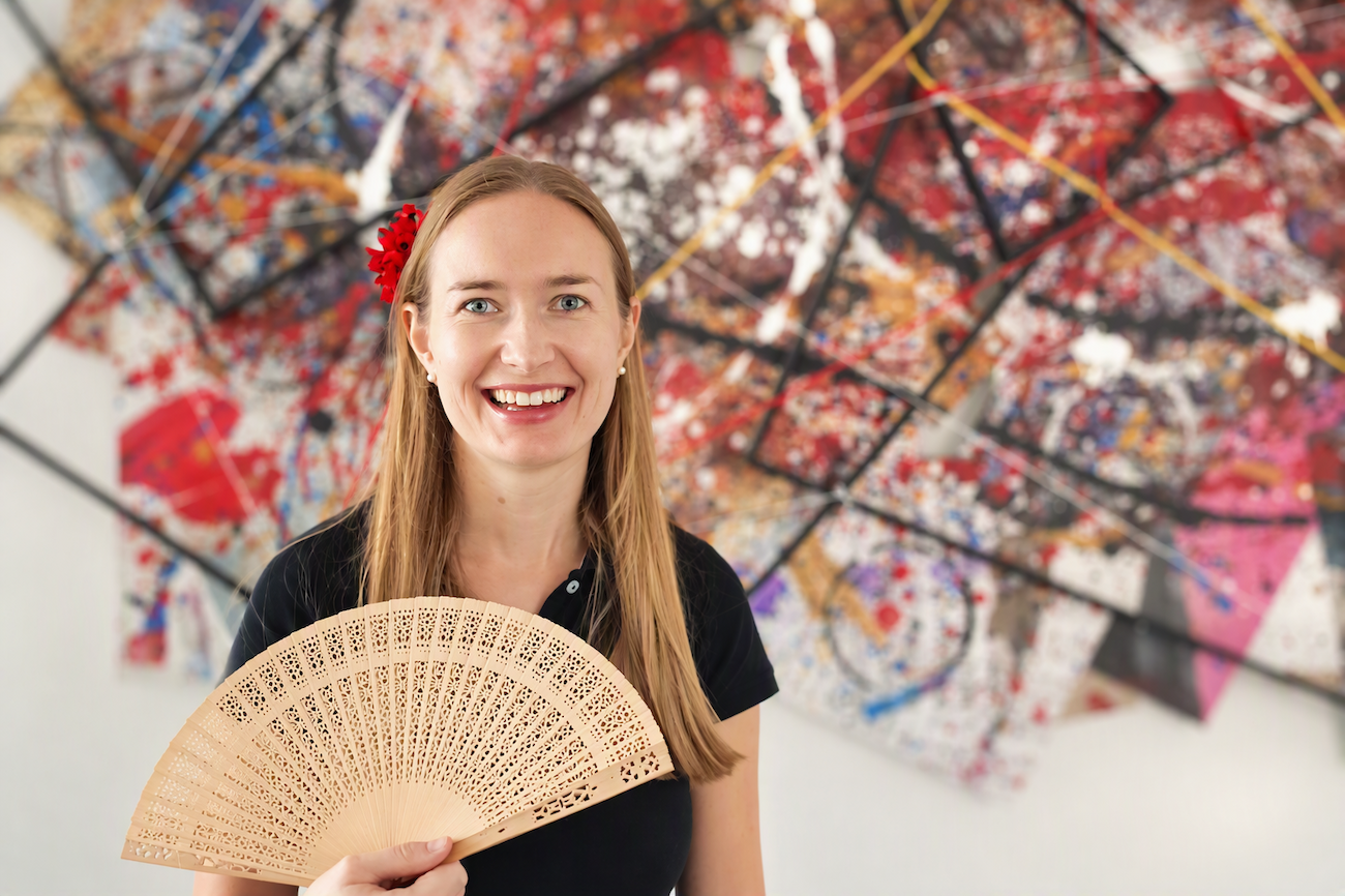 Eine lächelnde Frau mit einem Fächer steht vor einem bunten abstrakten Kunstwerk.