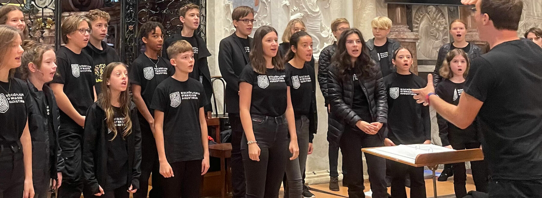 Ein Jugendchor singt in einer Kirche und folgt dabei den Anweisungen seines Dirigenten.