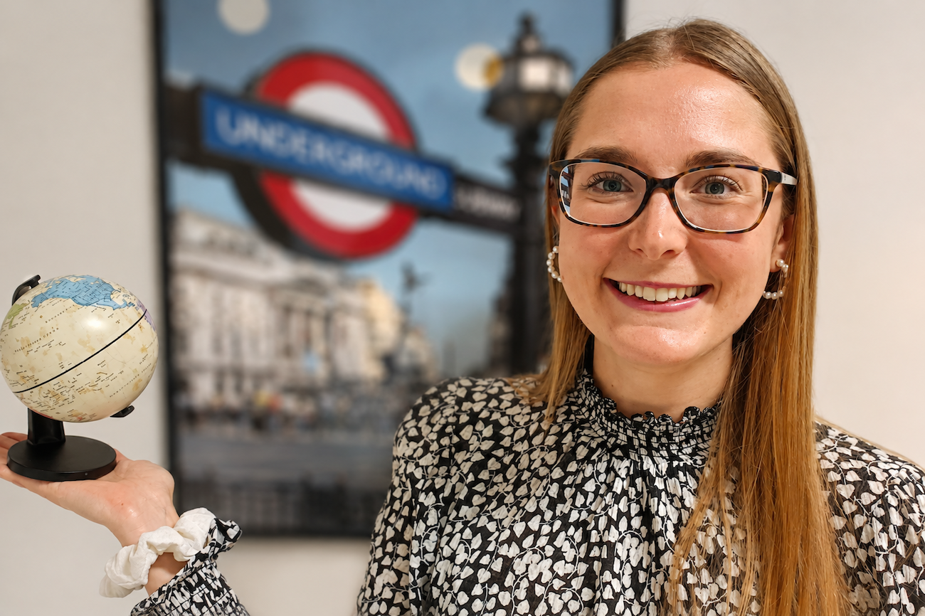 Lächelnde Frau mit einem kleinen Globus in der Hand vor einem Plakat der Londoner U-Bahn.