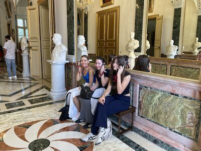 Drei junge Frauen sitzen auf einer Bank in einem Museum mit Marmorboden und Statuen.