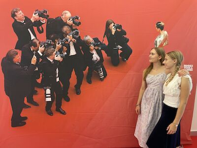 Zwei Frauen posieren vor einem roten Hintergrund, auf dem Fotografen aufgedruckt sind.