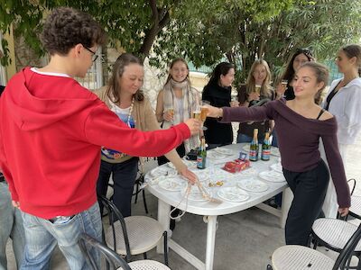 Eine Gruppe von Menschen, die lächelnd um einen weißen Tisch im Freien mit Essen und Getränken anstoßen.